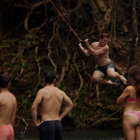 Person swings on a rope over water, watched by three others in swimsuits.