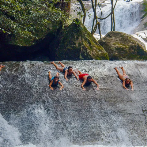 Five people enjoying a natural rock water slide at a waterfall in a forest.