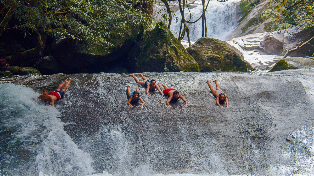 Five people sliding down a natural rocky waterfall in a forest.