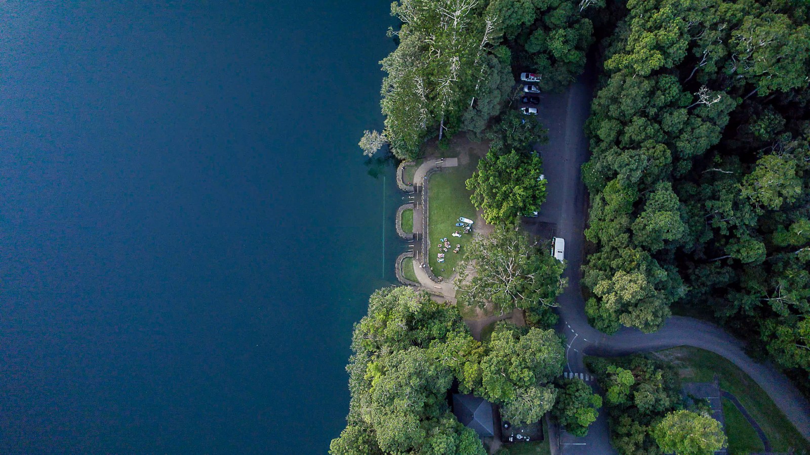 Aerial view of a lakeside park with grass, trees, and parked cars.