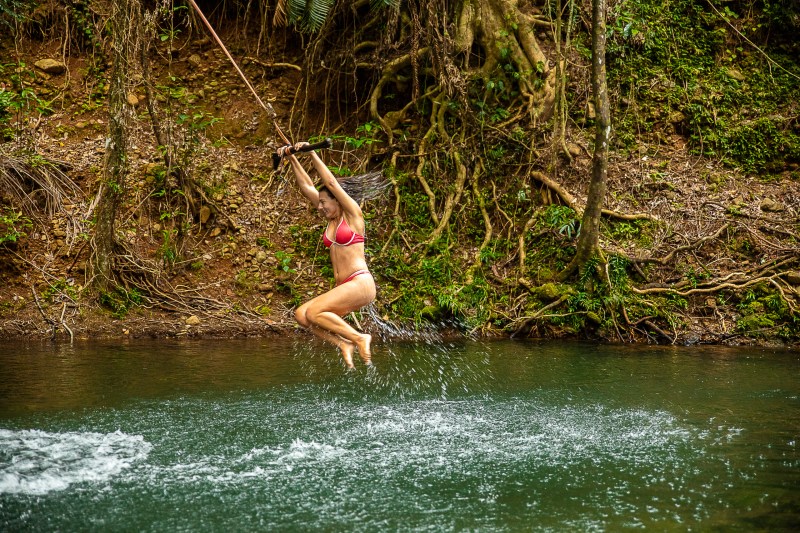 Person swinging on a rope over a river in the forest, wearing a red bikini.