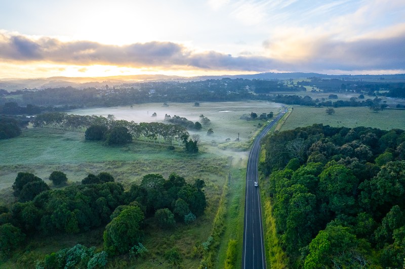 Aerial view of a misty rural landscape at sunrise with a road and scattered trees.