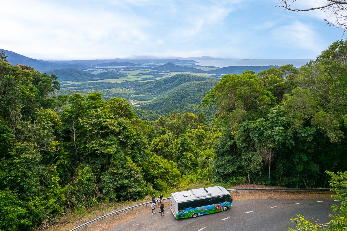 Van on winding road amidst lush green forest, overlooking valley and distant hills under a blue sky.