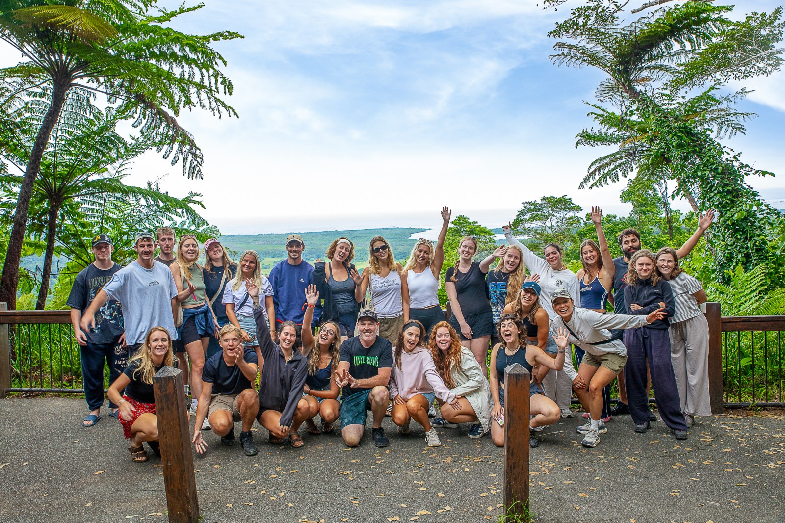 Group of people smiling and posing with arms raised in a scenic outdoor setting with trees and distant hills.